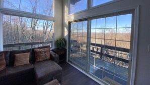 A room with a large sliding glass door leading to a deck, a dark-colored sofa with multiple cushions, and a small table with a plant. - Pocono Mountains Camelback Retreat Ski-in Ski-out (Tannersville)