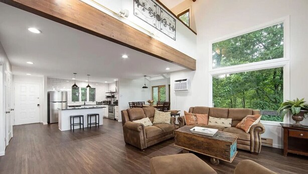 A spacious living area with a high ceiling, a kitchen in the background, and a large window offering a view of greenery. - Pocono Mountains Camelback Retreat Ski-in Ski-out (Tannersville)