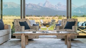 A room with a large window overlooking mountains, two sofas with pillows, a wooden table, and a small vase with flowers. - Basecamp Stays: Lux Retreat, Teton Views, 3 Acres! (Tetonia)