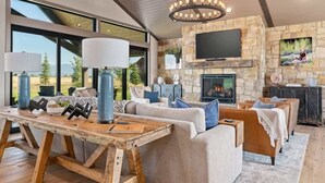 A modern living room with a stone fireplace, large windows, and a wooden dining table. - Basecamp Stays: Lux Retreat, Teton Views, 3 Acres! (Tetonia)