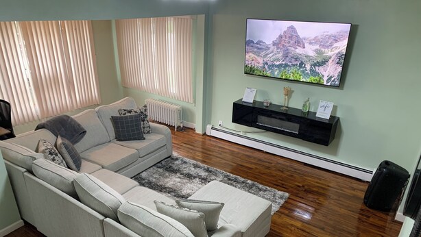 A living room with a sectional sofa, a flat-screen TV mounted on the wall, a radiator, and a small potted plant. - Charming 3-bedroom house in Queens with WiFi and AC (Queens)