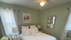 A bedroom with a bed, a nightstand, a lamp, a ceiling fan, a wall-mounted mirror, and a window with curtains. - Charming 3-bedroom house in Queens with WiFi and AC (Queens)