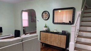 A living room with a sectional sofa, a wooden cabinet, a clock, and a staircase. - Charming 3-bedroom house in Queens with WiFi and AC (Queens)