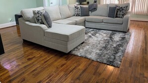 A living room with a sectional sofa, a rug, wooden flooring, a door, and a wall-mounted TV. - Charming 3-bedroom house in Queens with WiFi and AC (Queens)