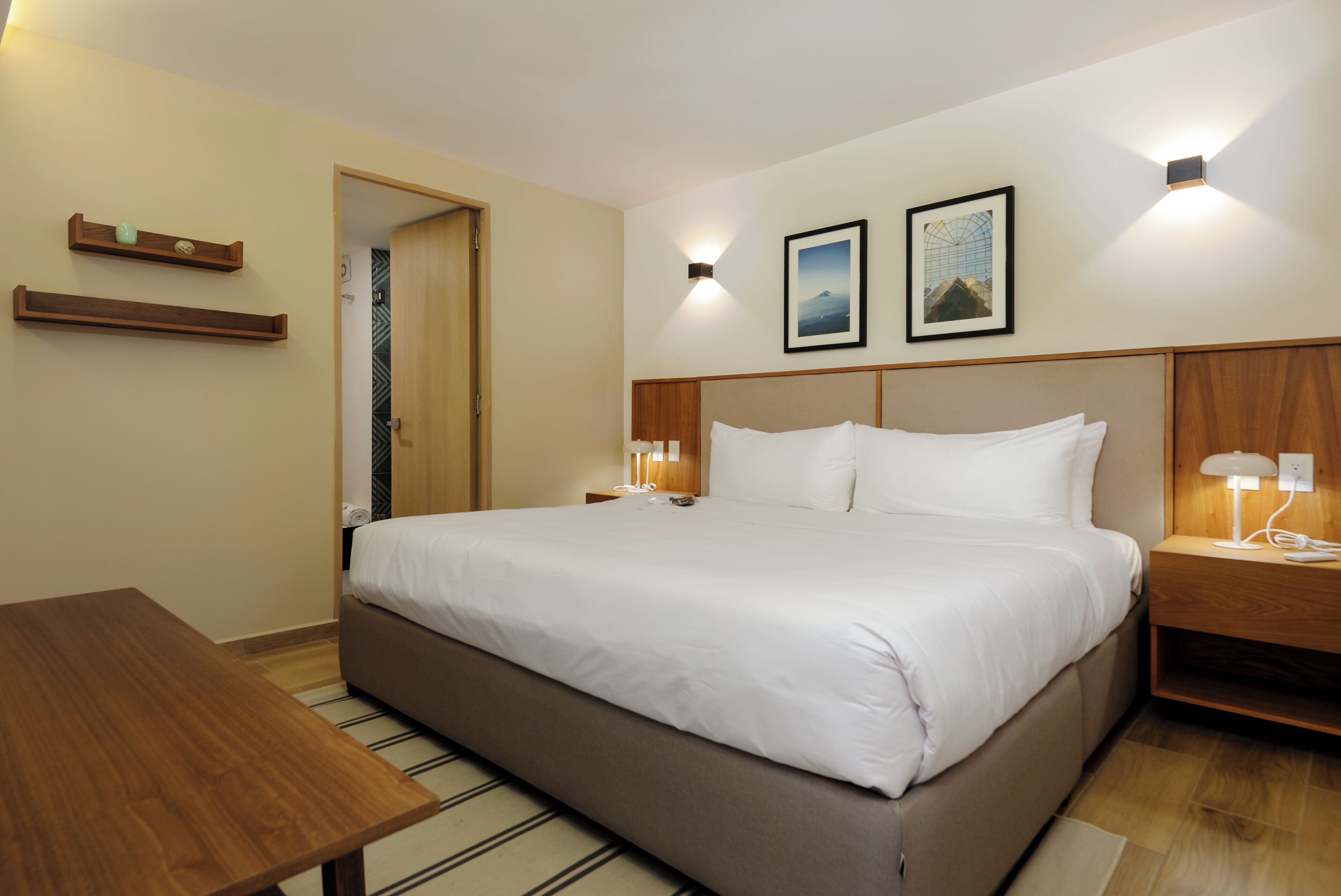 Habitación de hotel con una cama grande, mesitas de noche, una mesita auxiliar de madera, una mesa de madera y dos cuadros colgados en la pared.