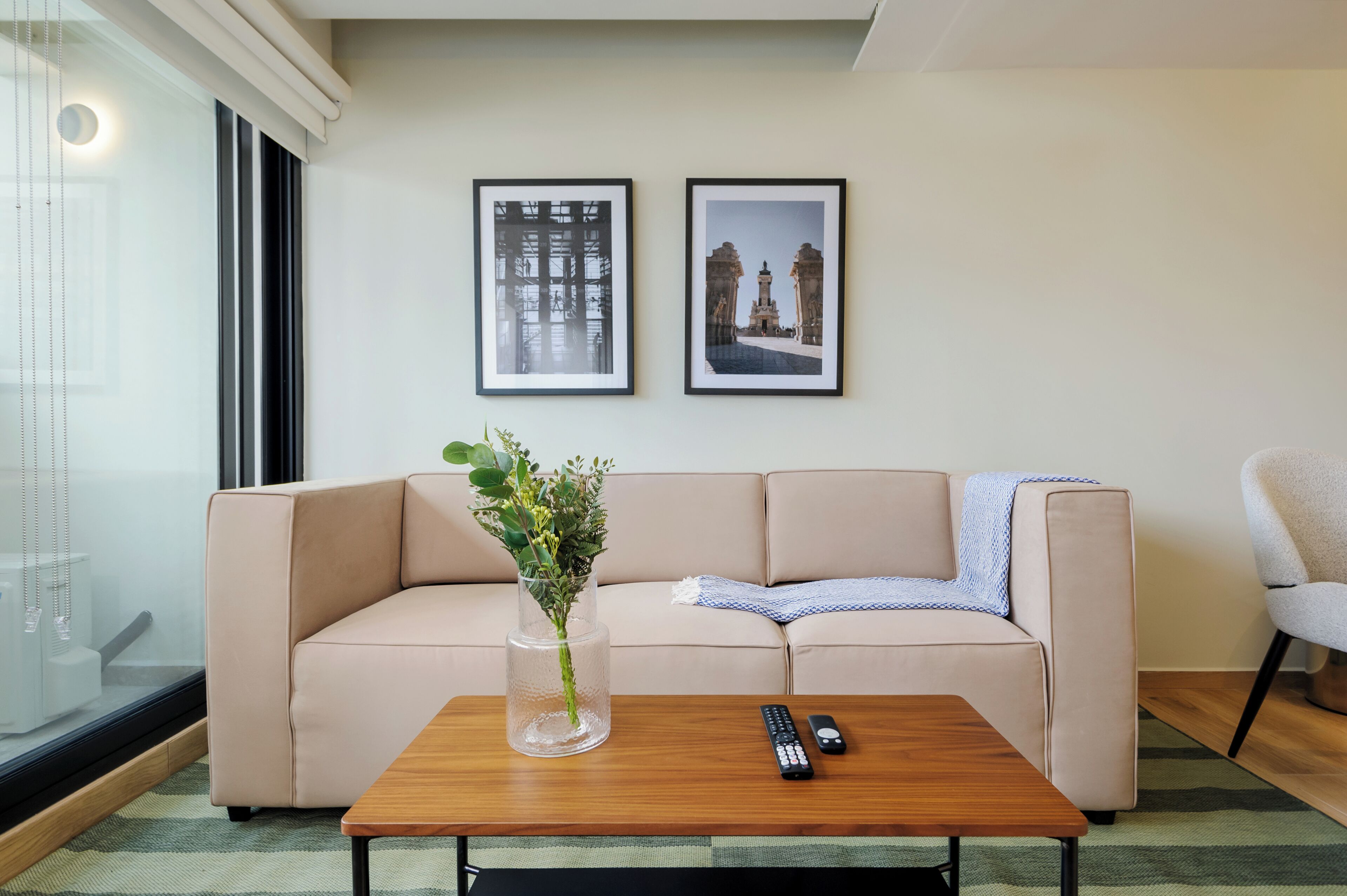 A living room with a beige sofa, a wooden coffee table, two framed pictures on the wall, a vase with green plants, and a remote control on the table.