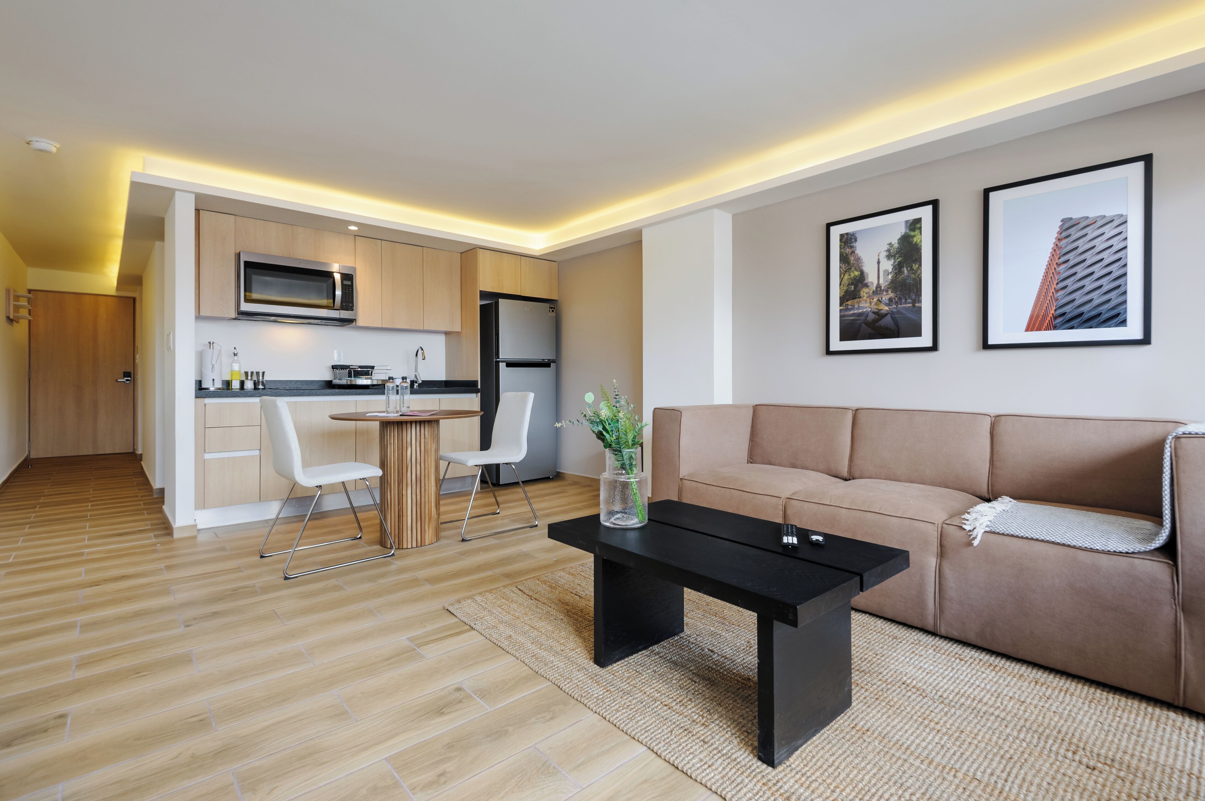 En moderne lejlighed med et køkken, spiseområde og opholdstue. Den har trægulv, et sort sofabord, en beige sofa og billeder i rammer på væggen.