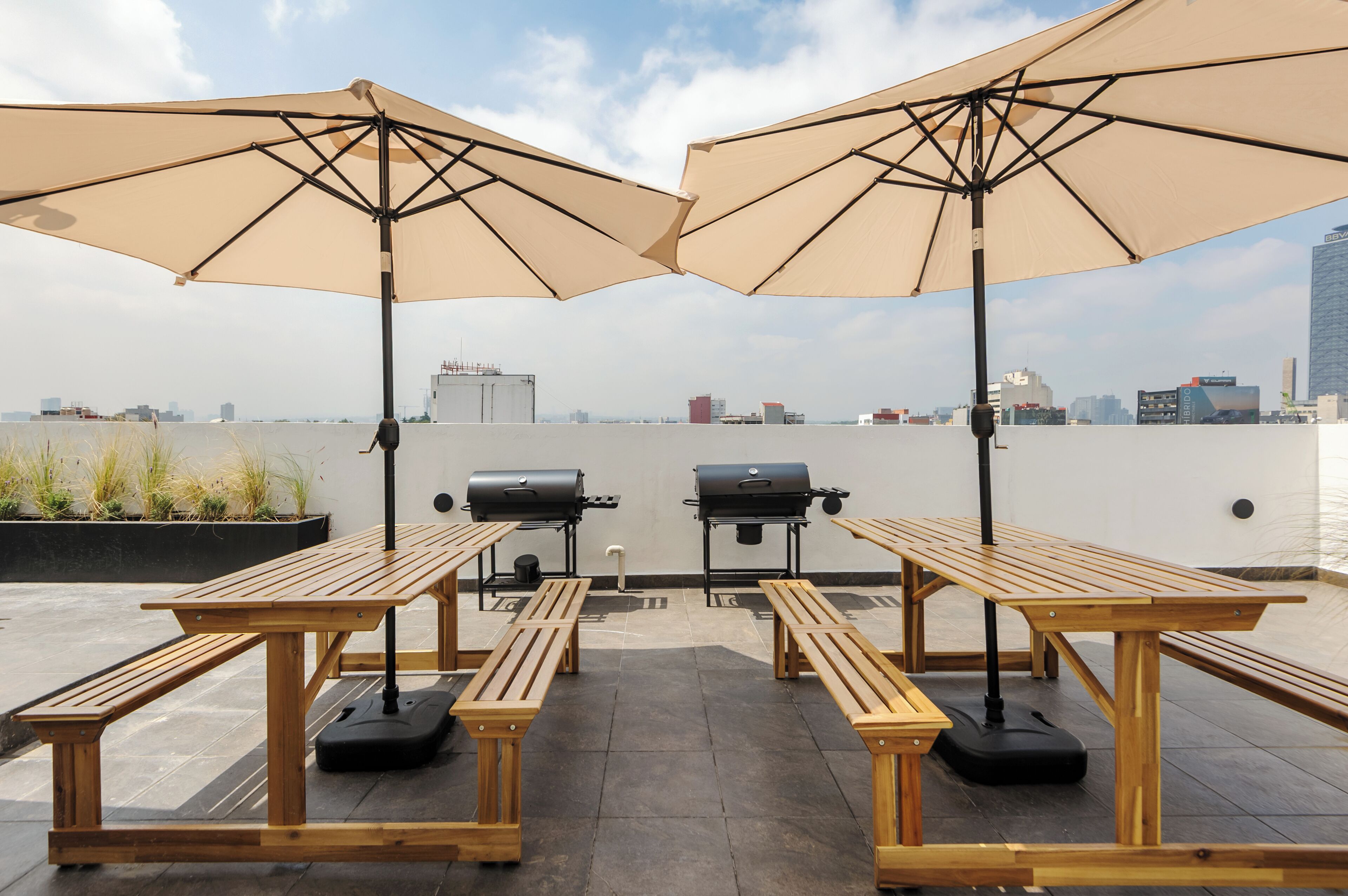 An outdoor patio with wooden tables and benches, two barbecue grills, and large umbrellas.