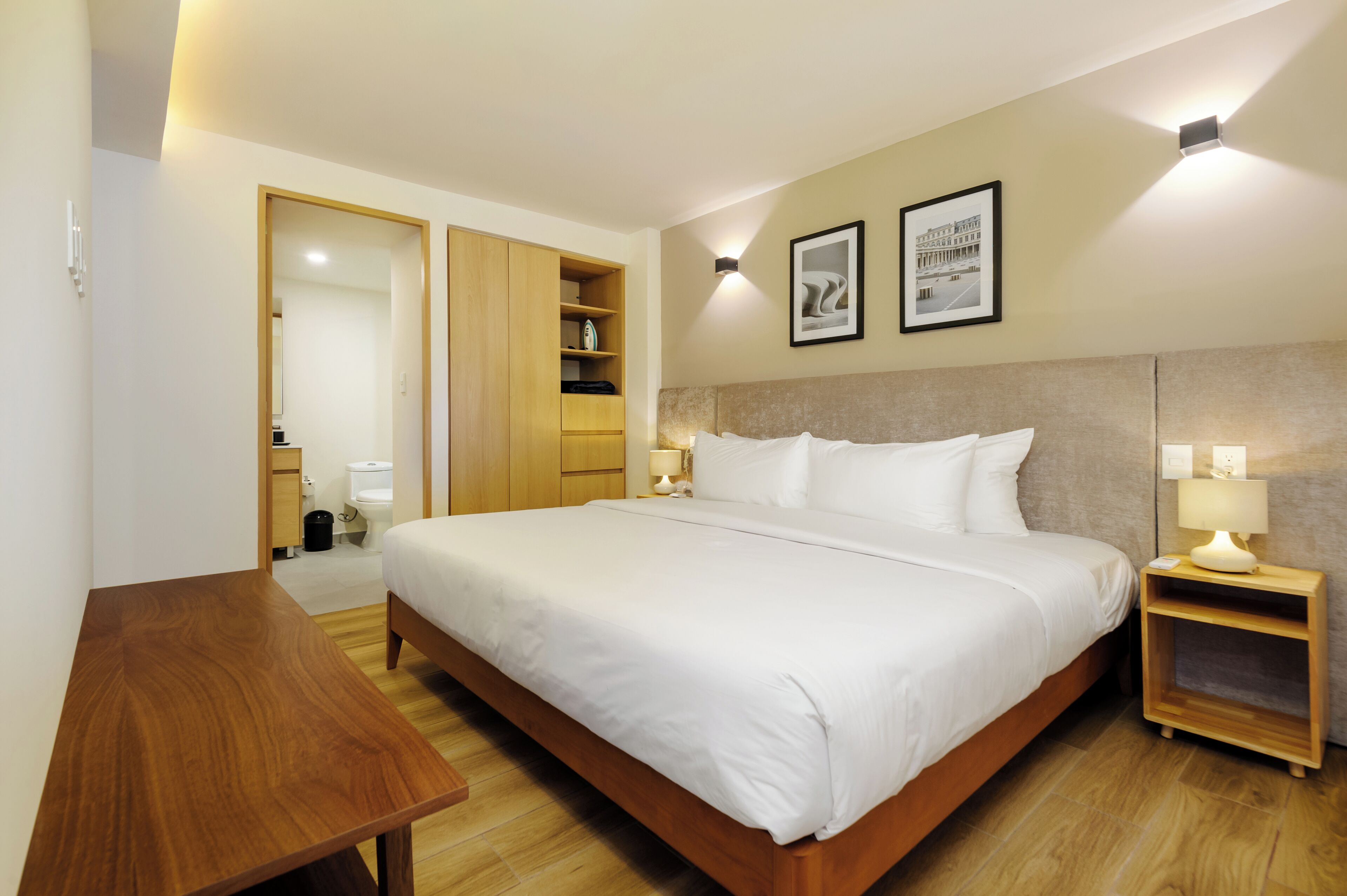 A hotel room with a large bed, wooden nightstands, a wooden desk, a wardrobe, and a bathroom visible through an open door.