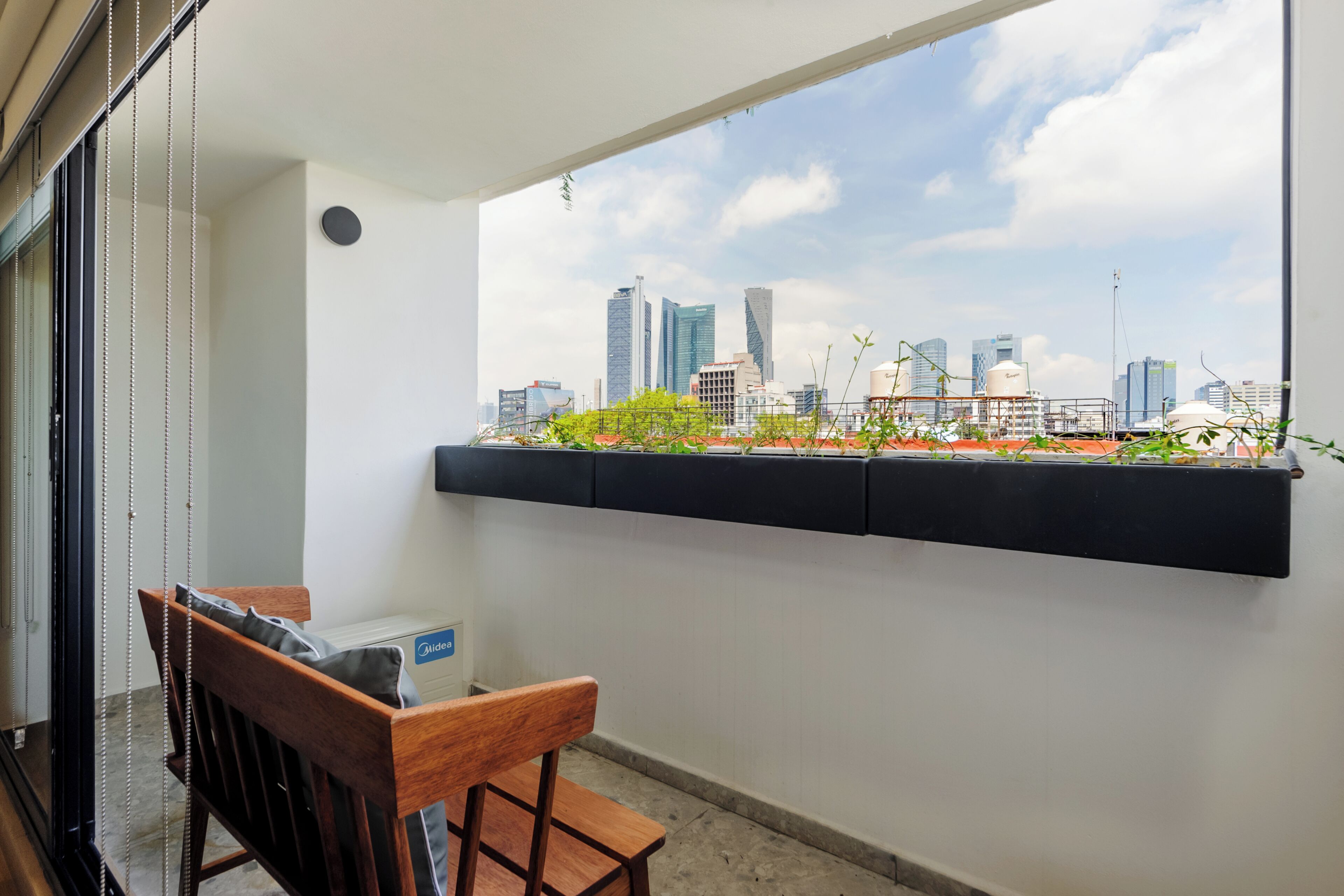 Un balcon avec vue sur la ville, une chaise en bois et un pot noir contenant des plantes.