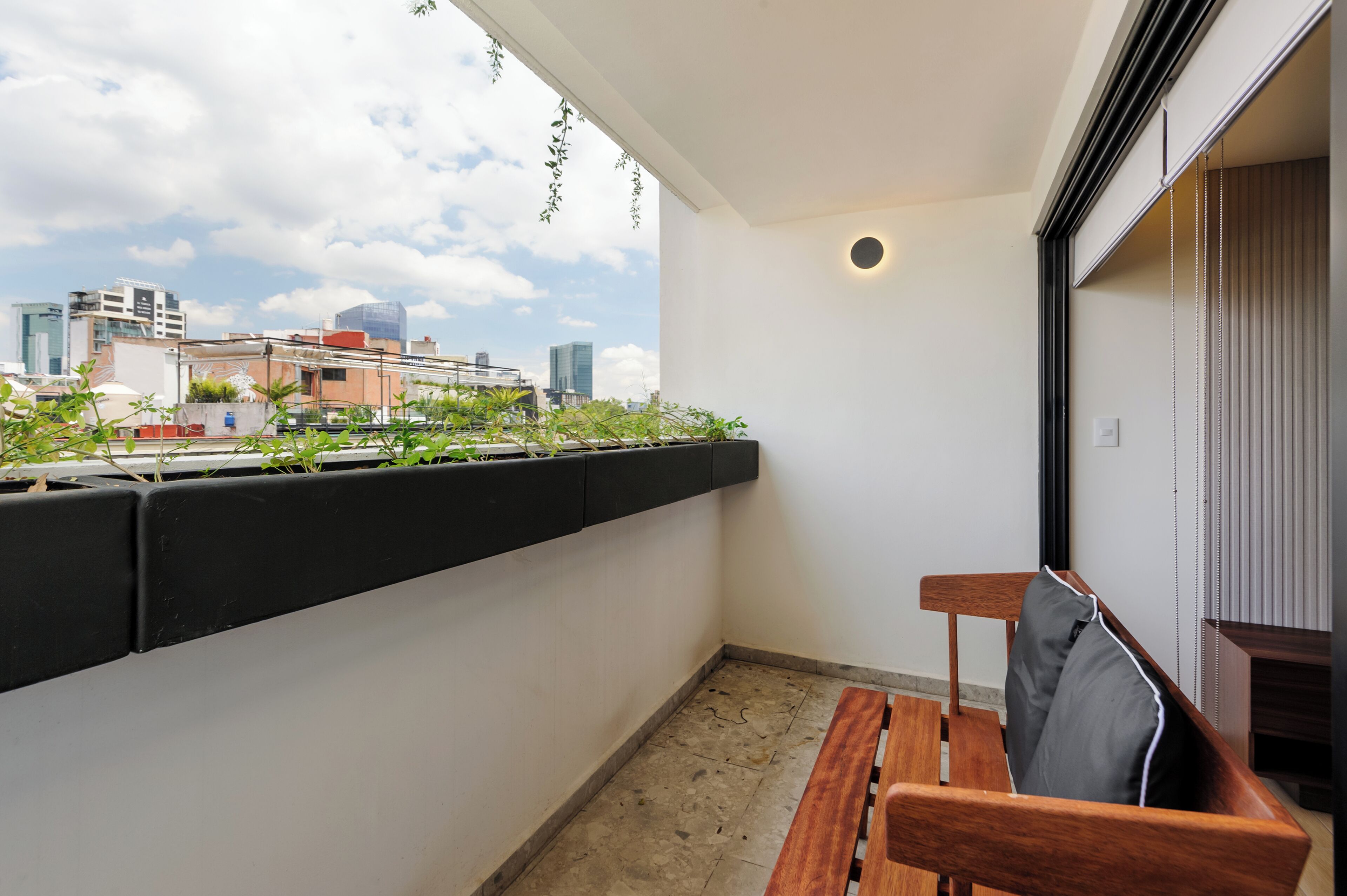 Un balcon avec un banc en bois, un pot noir contenant des plantes vertes et une vue sur la ville.