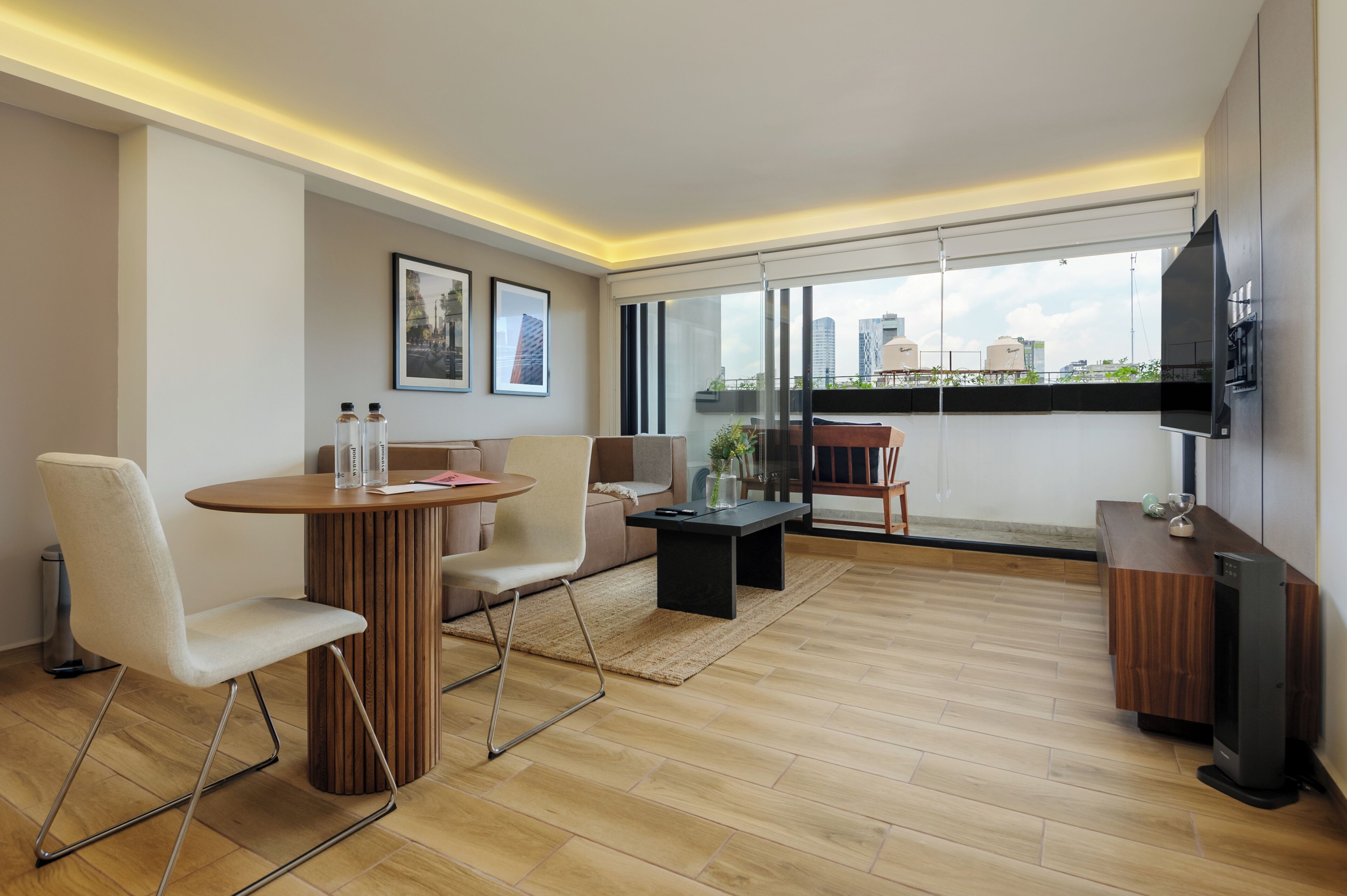 Uma sala de estar moderna com piso de madeira, uma mesa redonda, duas cadeiras, um sofá, uma TV e uma grande janela com vista para a cidade.