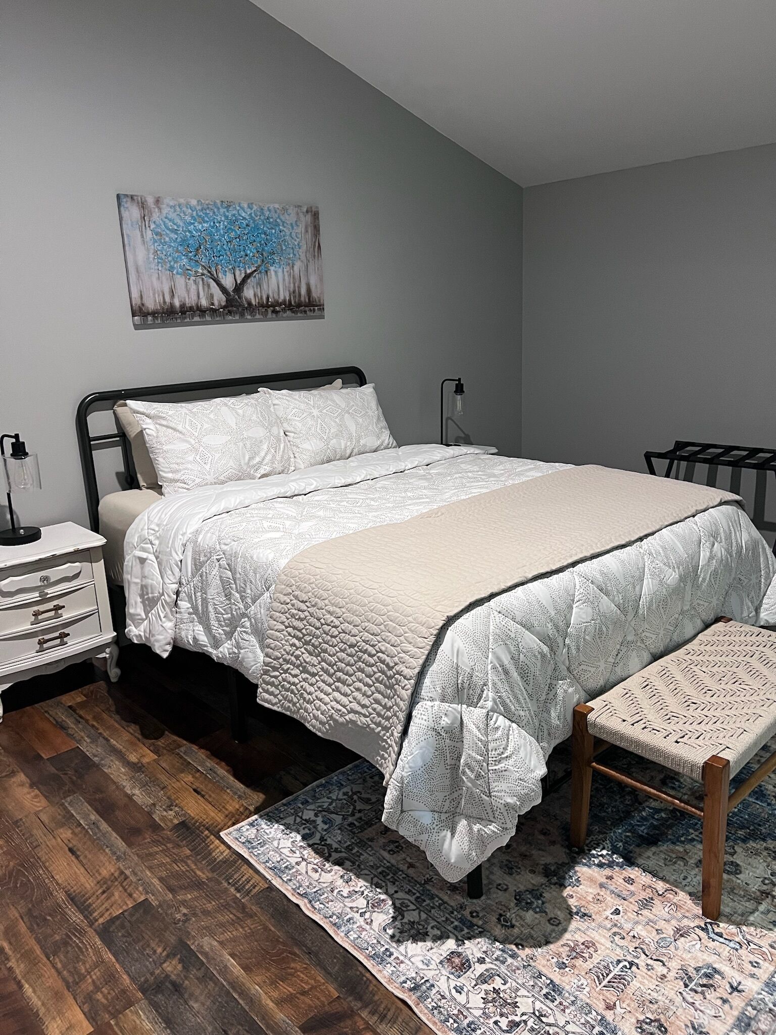 A bedroom with a bed, bedside tables, a nightstand, a chair, a rug, and a painting on the wall.