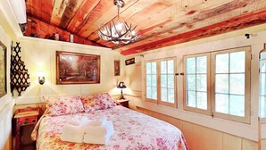Une pièce chaleureuse avec un plafond en bois, un lit recouvert d’une literie à motifs floraux, un lustre et de grandes fenêtres.