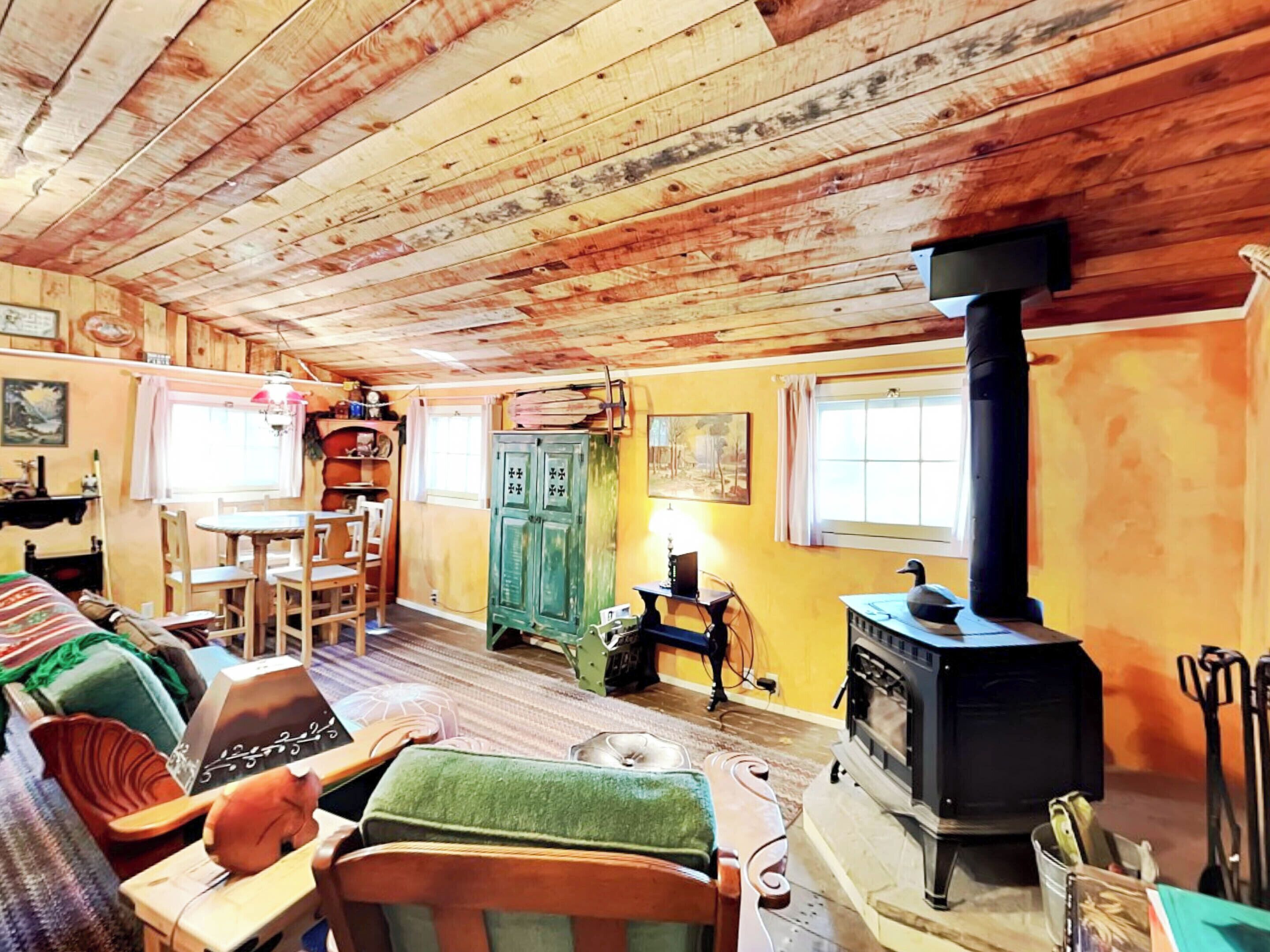 A cozy cabin interior with wooden walls and ceiling, a dining area with chairs and a table, a green cabinet, and a wood-burning stove.