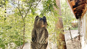 Une sculpture en bois représentant un ours, dans un décor naturel avec des arbres et un bâtiment en arrière-plan.
