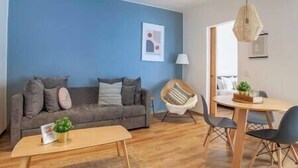 Een woonkamer met een blauwe wand, een grijze bank met kussens, een houten salontafel, een witte stoel en een eettafel met stoelen.