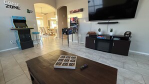 Un salon comprenant un téléviseur, une table basse avec un jeu de société, une console de jeux et un meuble TV agrémenté de plantes et d’une horloge.