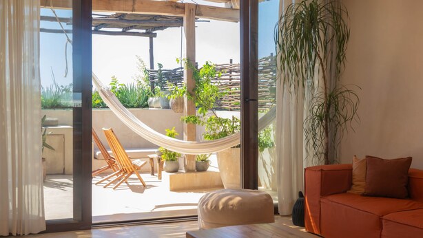 Un salon moderne avec un canapé, un banc en bois, un hamac et une vue sur une terrasse extérieure agrémentée de plantes et d’une clôture en bois.