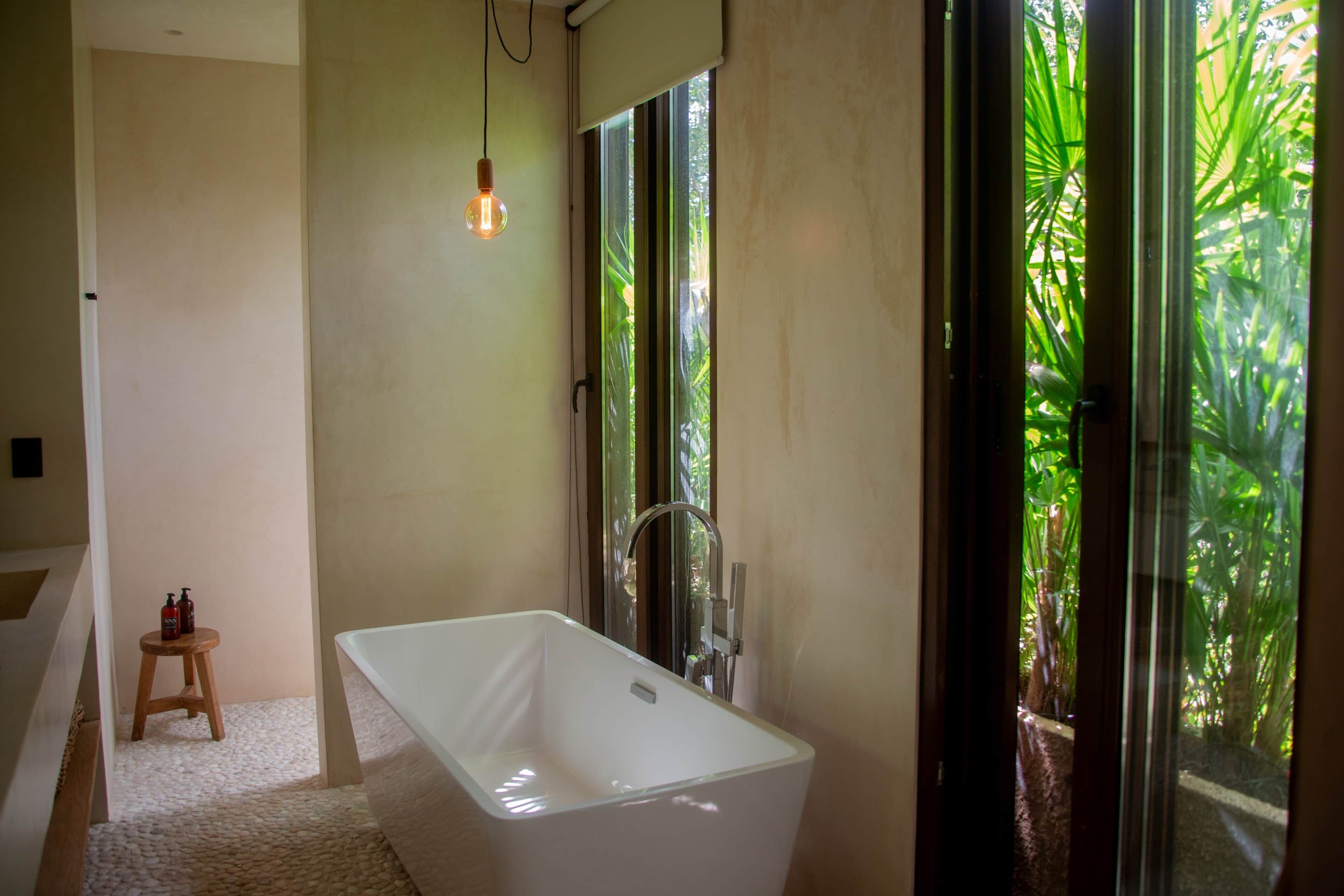 Un baño moderno con una bañera independiente, un taburete de madera y un amplio ventanal que ofrece vistas de abundante vegetación.