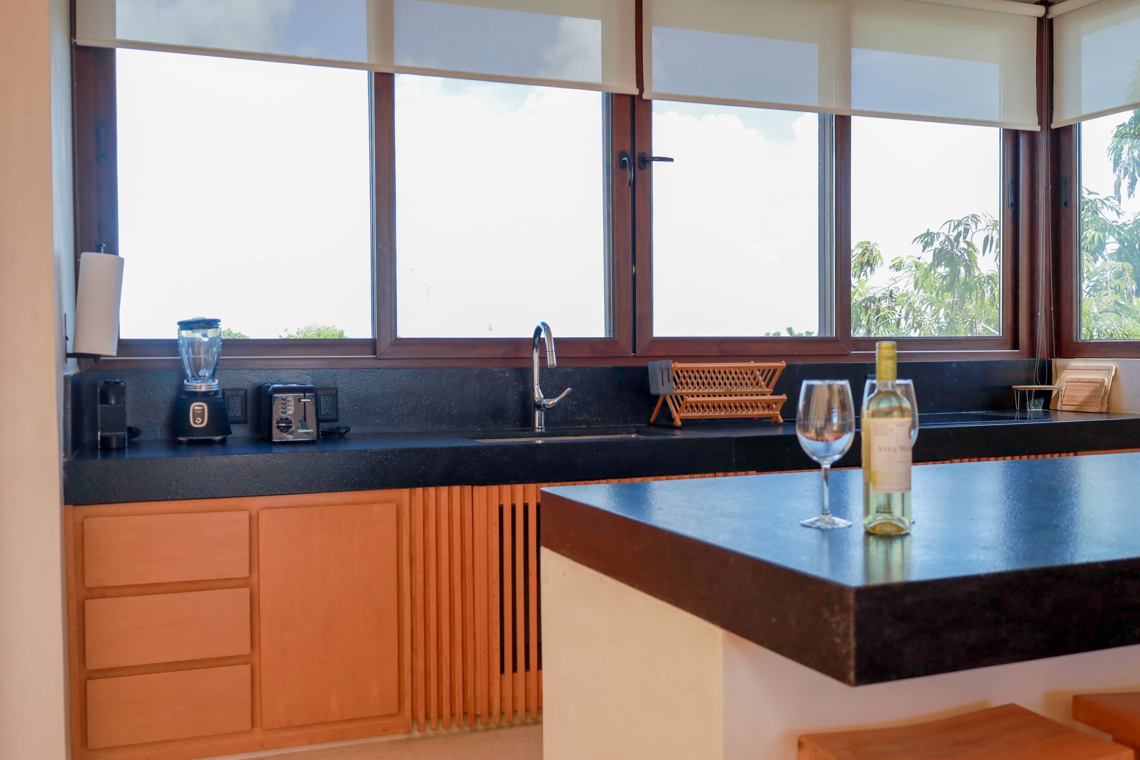 Una cocina con encimera negra, armarios de madera y una ventana con persianas, además de una copa de vino.