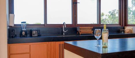 Una cocina con encimera negra, armarios de madera y una ventana con persianas, además de una copa de vino.