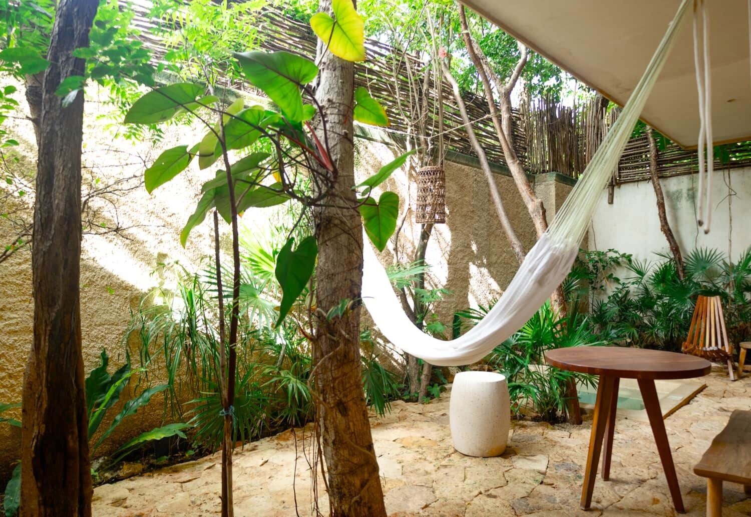 A hammock, a round table, and a bench in a garden with trees and plants.