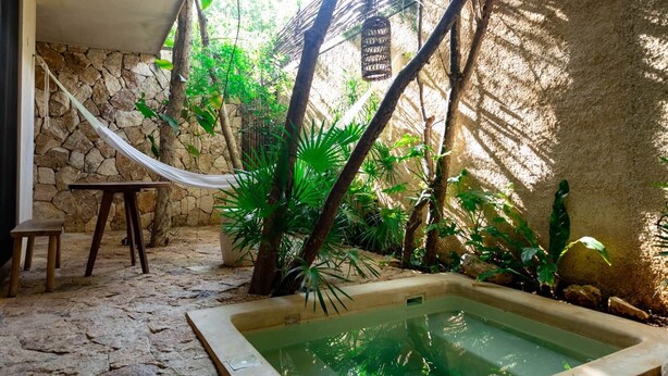 A stone-walled room with a hammock, a small table, and a hot tub surrounded by tropical plants. - Room with Private Pool and Garden |• TEVA 2B (Tulum)