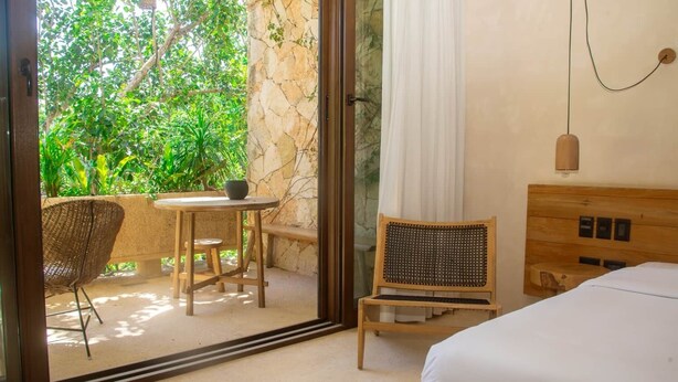 Habitación de hotel con una cama, una mesita con sillas, un sillón de mimbre y vistas a un follaje exuberante a través de un ventanal.