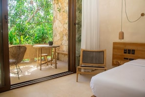 A hotel room with a bed, a small table with chairs, a wicker chair, and a view of lush greenery through a glass door. - Room with Private Jungle Balcony |• TEVA 6B (Tulum)