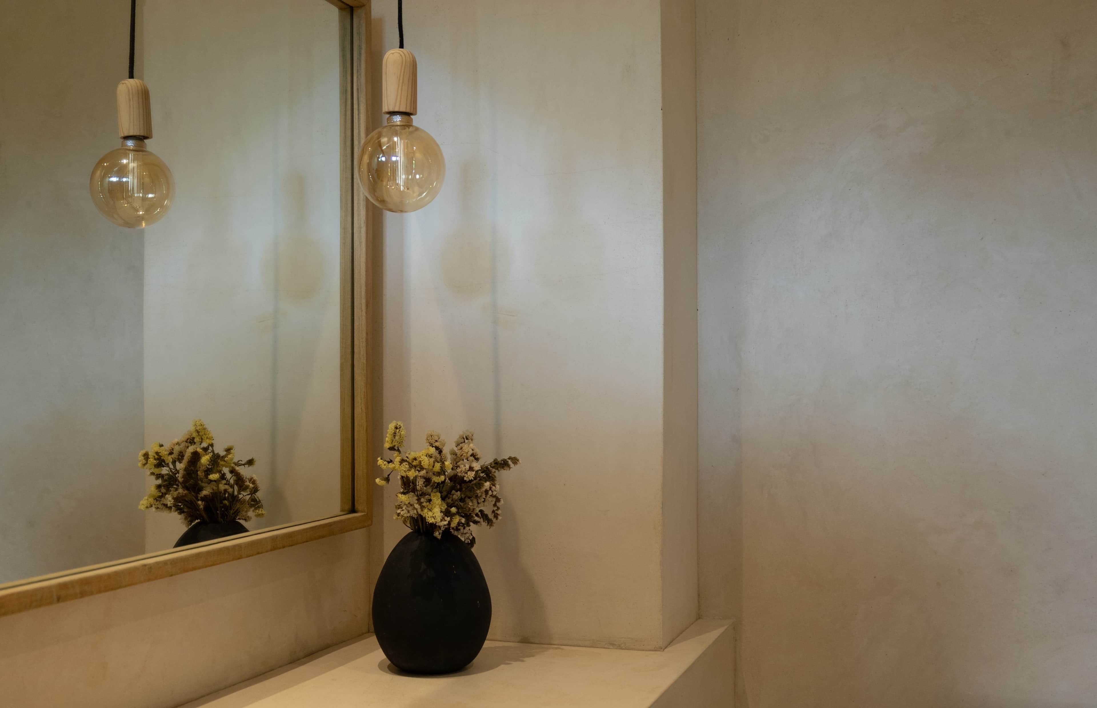 Casa de banho com espelho, duas lâmpadas pendentes e um vaso com flores secas numa prateleira branca.