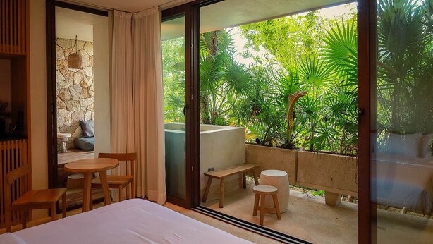 A modern hotel room with a large bed, a wooden table, and a bench. The room has a view of lush greenery outside through large glass doors. - Room with Private Terrace and Pool |• TEVA 4B (Tulum)