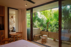 Una habitació d'hotel moderna amb un llit gran, una taula de fusta i una panxa. La habitació té vista a una vegetació abundant a l'exterior a través de grans portes de vidre.