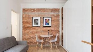 A small room with a brick wall, two framed pictures, a grey sofa, a round table with two chairs, and a wooden floor. - Cozy · Cozy apartment great location (Mexico City)
