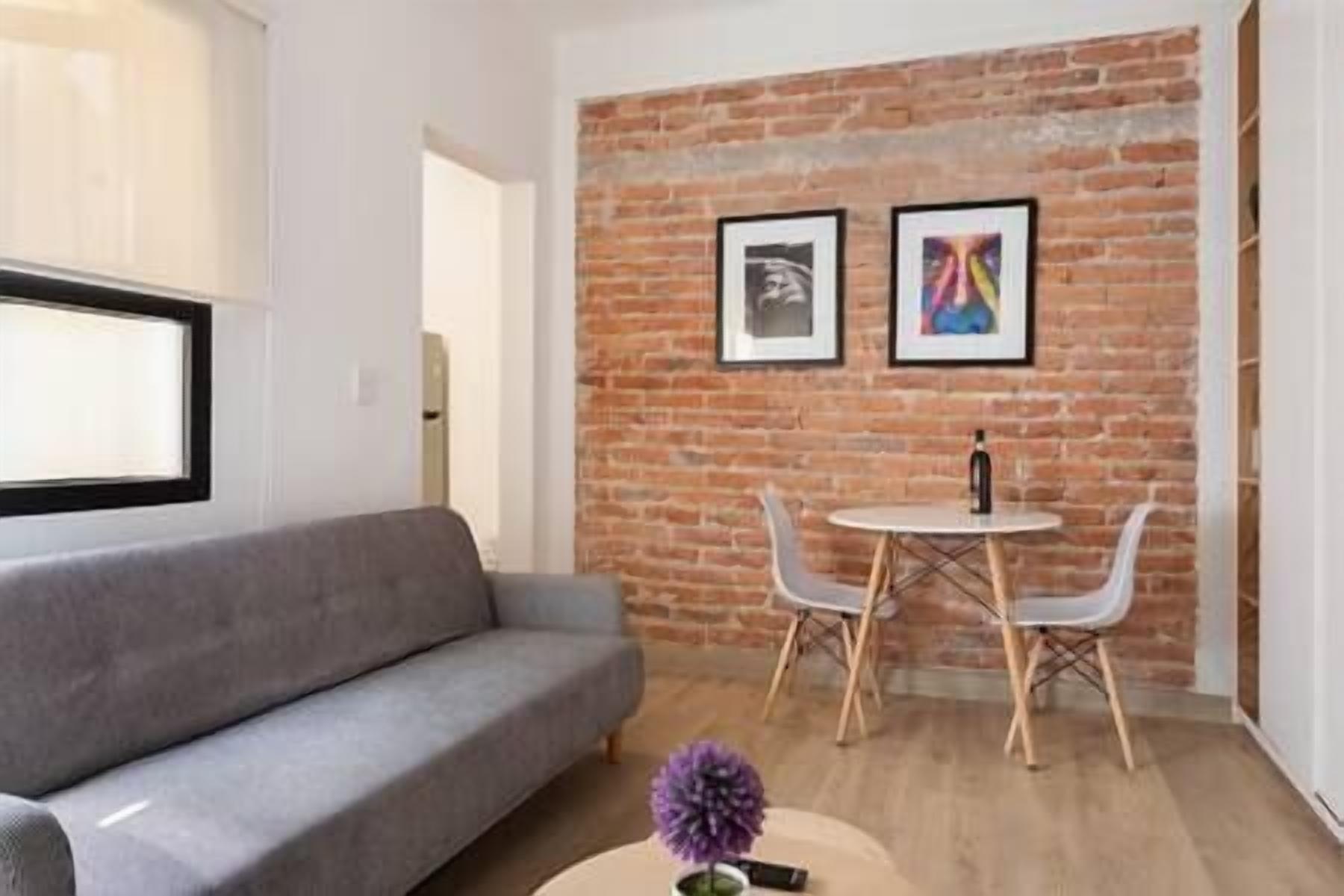 A modern living room with a grey sofa, a small round table with two chairs, and a brick wall with two framed pictures.