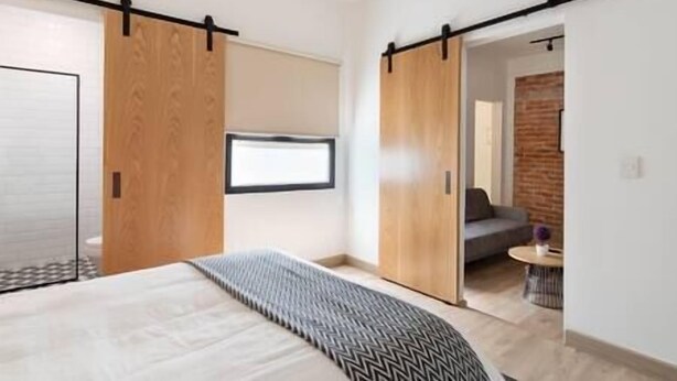 A modern bedroom with a large bed, a sliding door, and a view into another room with a sofa and a brick wall. - Cozy · Cozy apartment great location (Mexico City)