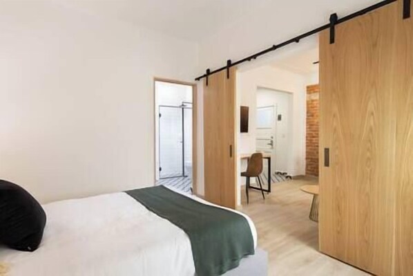 A modern hotel room with a bed, a desk, and a chair. There is a sliding door leading to another room.