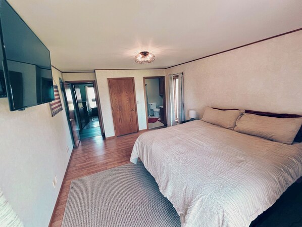 Une chambre à coucher avec un grand lit, un plancher en bois, un luminaire au plafond et une porte donnant sur une autre pièce.