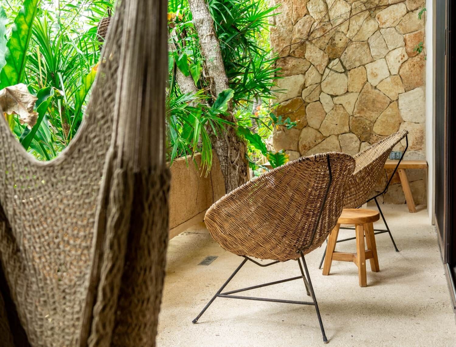A wicker chair with metal legs, a wooden stool, and a stone wall in a room with a concrete floor and green plants.