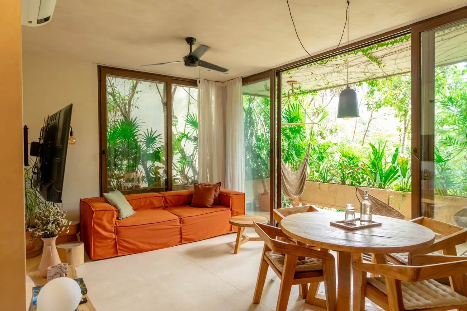 A modern living room with a sofa, dining table, and chairs. Large windows offer a view of greenery outside.