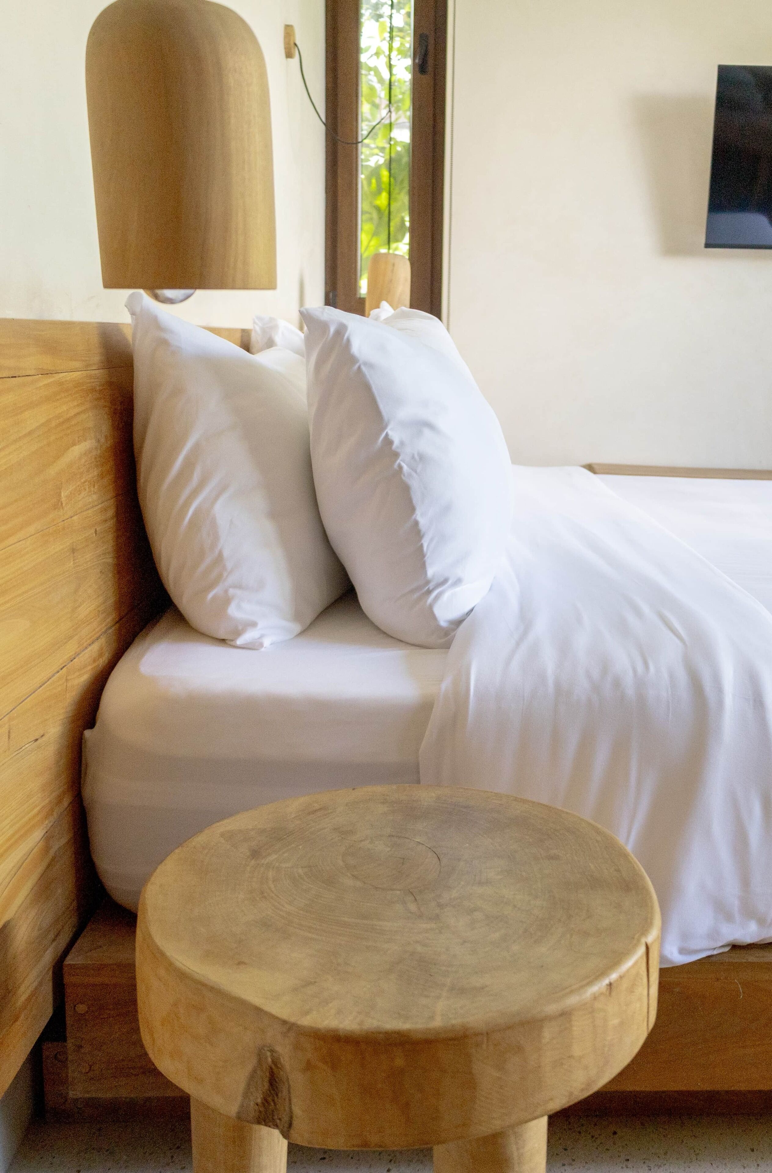Una cama bien tendida con ropa de cama blanca, un cabecero de madera y un taburete redondo también de madera en una habitación de diseño sencillo y limpio.