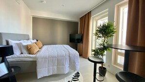 A modern hotel room with a bed, a desk, a chair, a standing desk, a potted plant, and a large window with curtains. - Borg Housing Travel Center (Pori)