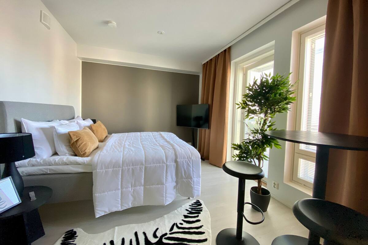 Ein modernes Hotelzimmer mit Bett, Schreibtisch, Stuhl, höhenverstellbarem Tisch, Zimmerpflanze und einem großen Fenster mit Vorhängen.
