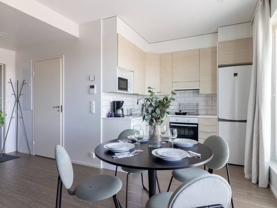 A modern kitchen with a dining area, featuring a round table set for four, a coffee machine, and a refrigerator.