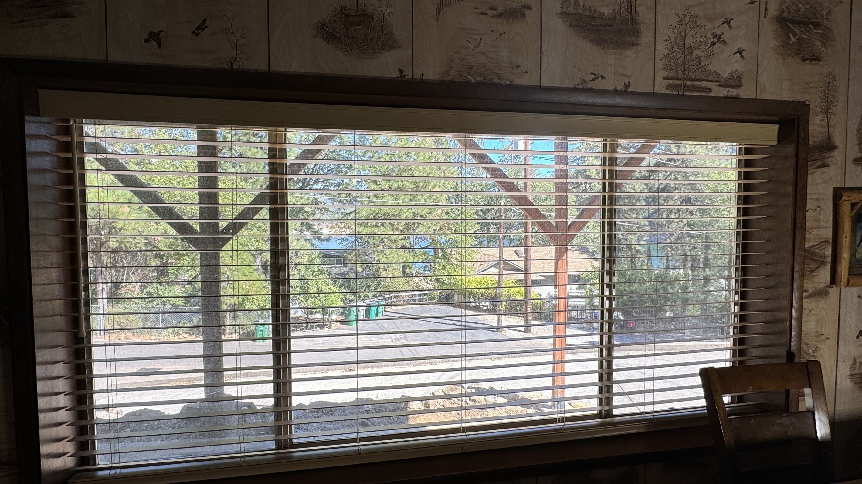 Een kamer met een groot raam met horizontale zonwering, een houten stoel en een muur met een muurschildering.