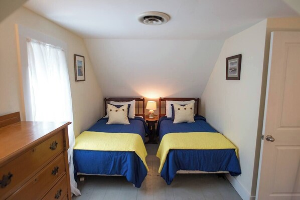 A small, single-room accommodation with two beds, each with a blue and yellow bedspread, a wooden dresser, and a door on the right.