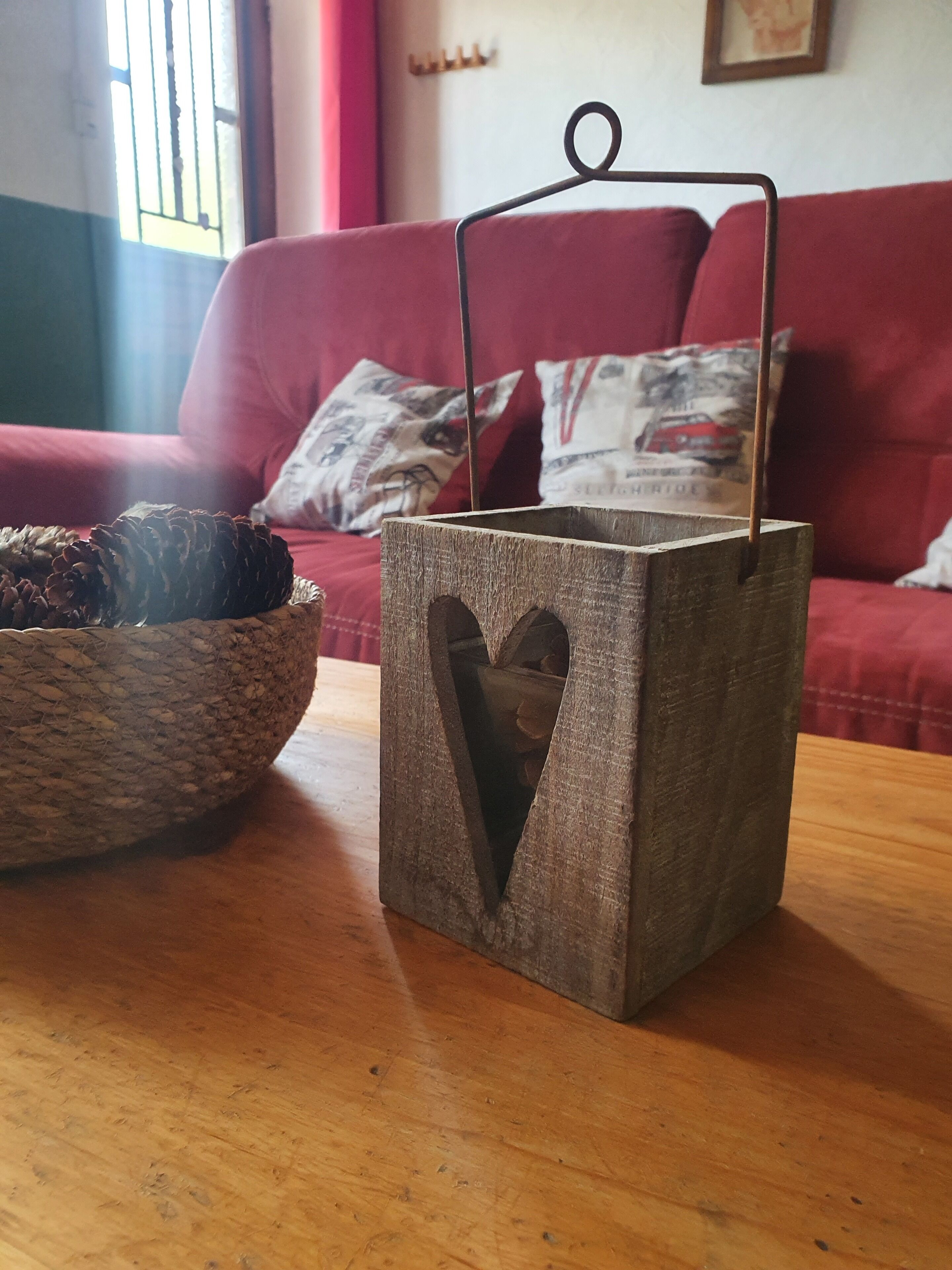 Eine rustikale Holzlanterne mit Herzförmigem Ausschnitt auf einem Holztisch, ein geflochtener Korb mit Kiefernzapfen und eine rote Couch mit gemusterten Kissen im Hintergrund.