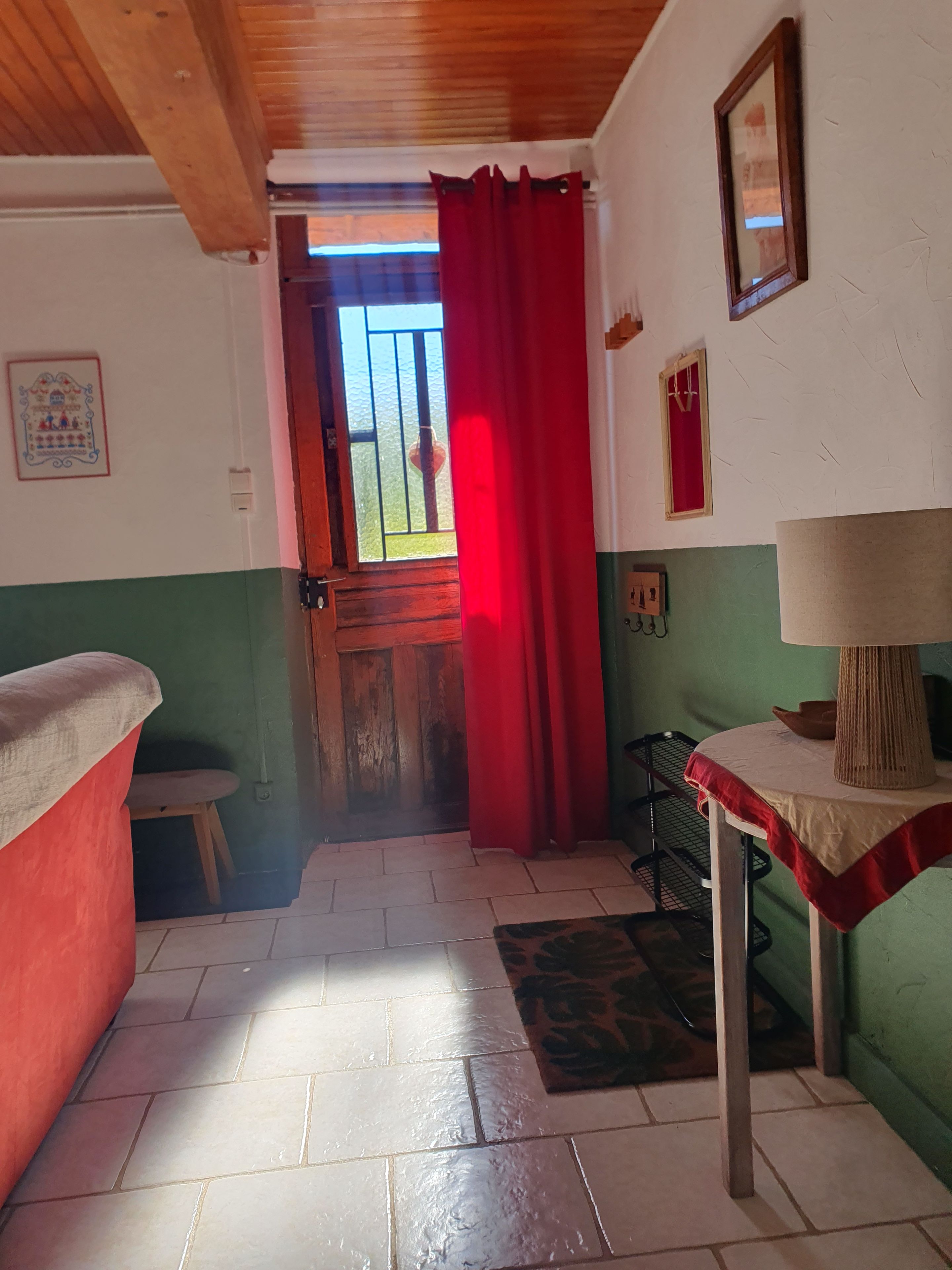 Una habitación con techo de madera, una cortina roja, un taburete pequeño, una mesa con una lámpara y una pared verde.