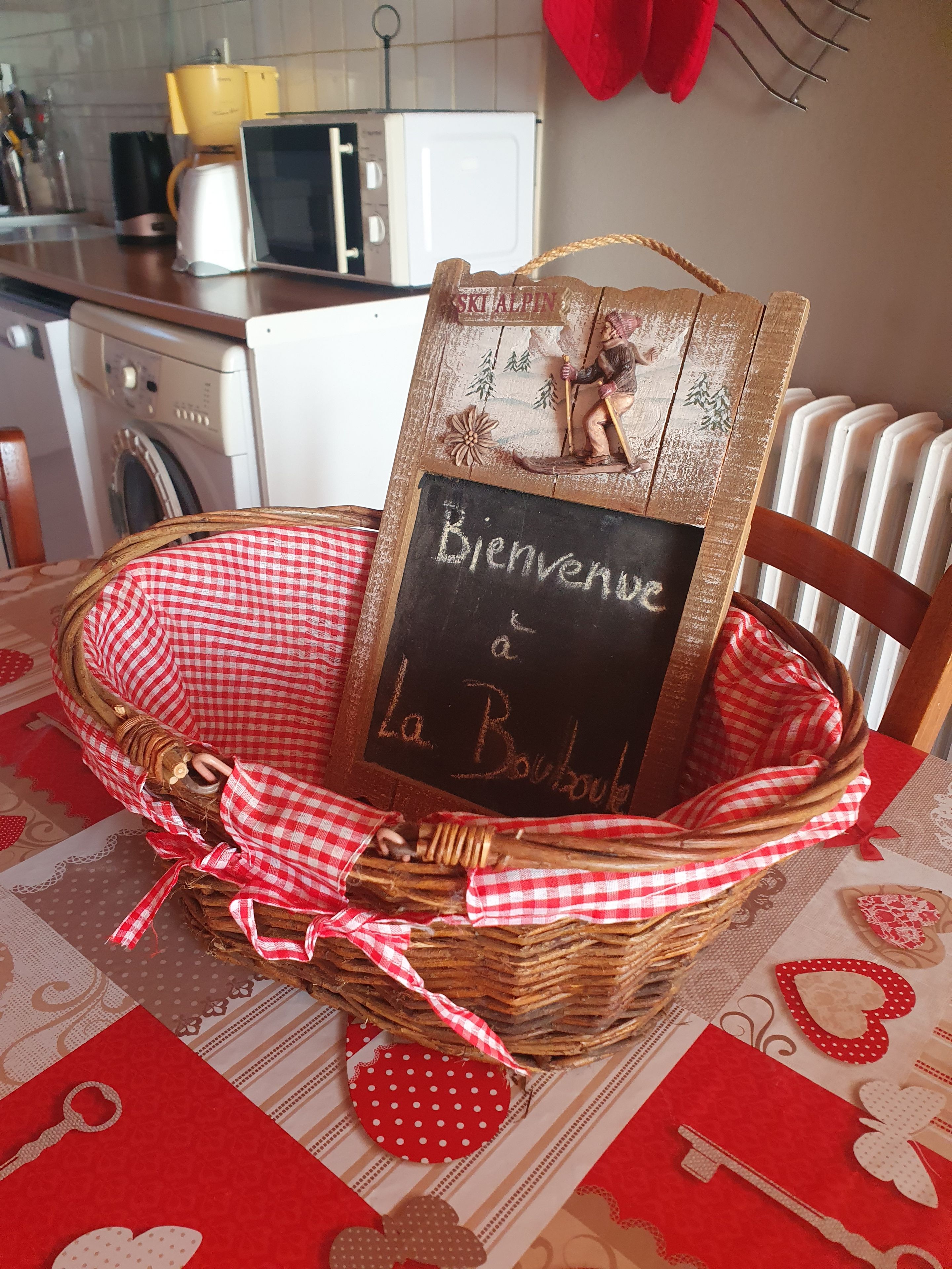 Un cesto di vimini con una tovaglia a quadretti rossi e bianchi, contenente un cartello di legno raffigurante uno sciatore e la scritta 'Bienvenue à La Bouboule'.