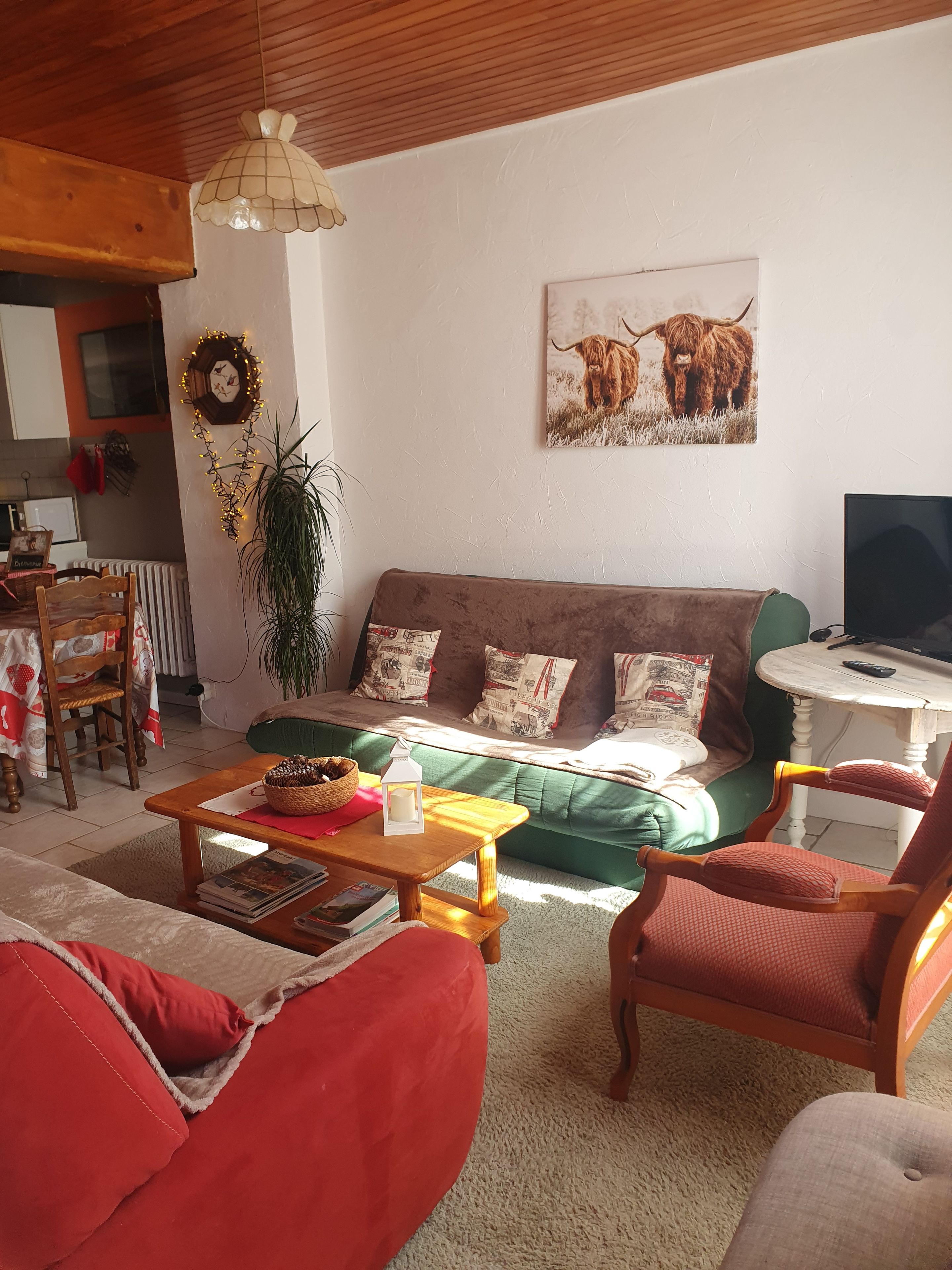 Una sala de estar acogedora con un sofá verde, una mesa de centro de madera y sillones rojos. Un televisor está sobre una mesita auxiliar, y hay un reloj de pared con iluminación.
